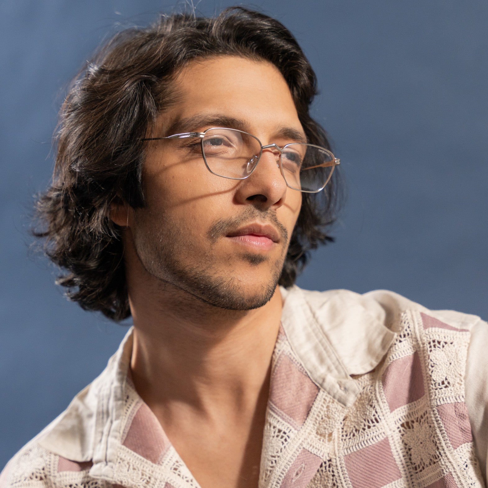 Andreas Edition Silver Anti Blue Light Trapezoid Sunglasses – Sunglassic.com. Male model wearing minimal silver metal-frame trapezoid glasses with clear blue light protection lenses, styled with a vintage crochet-patterned shirt against a soft blue background.