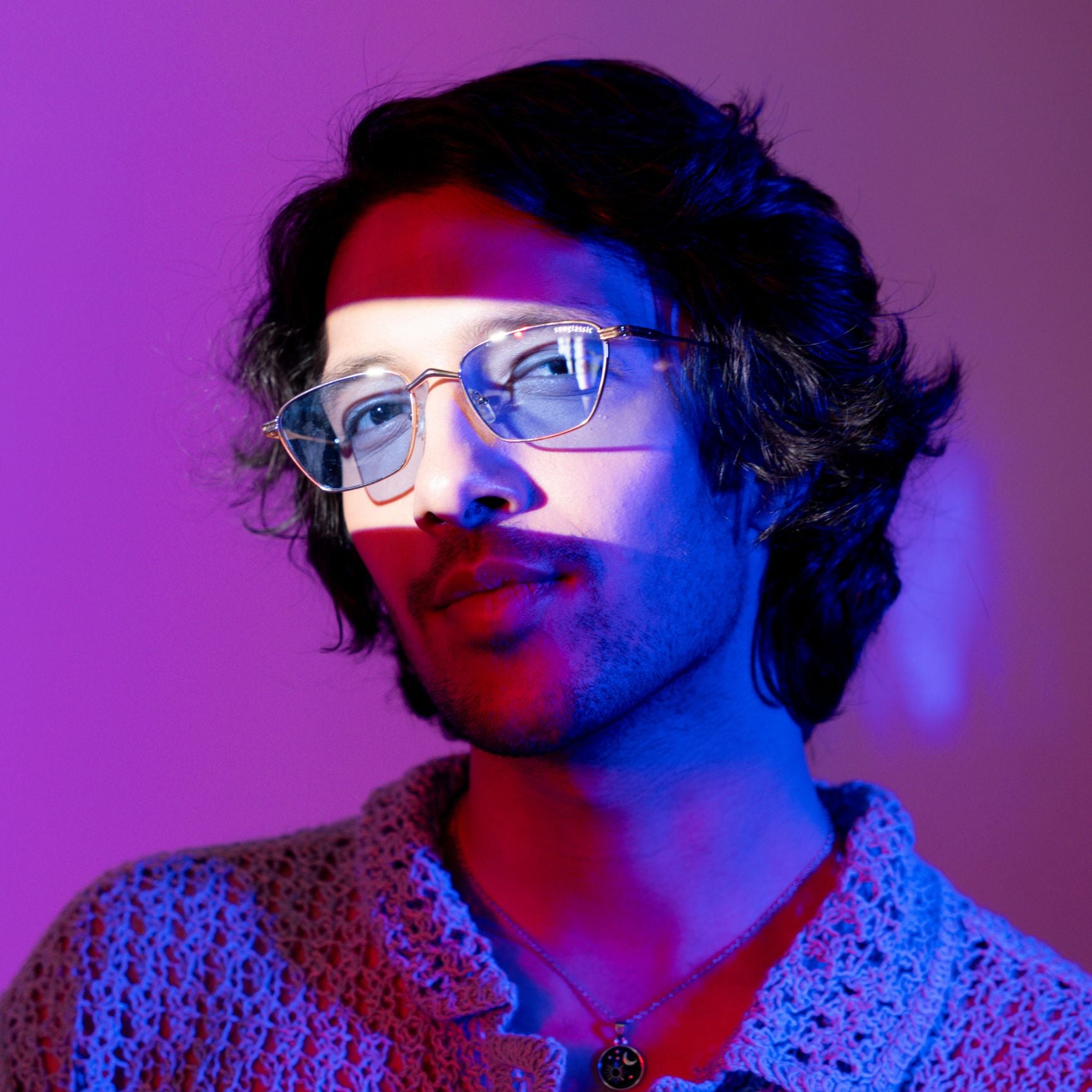 Andreas Gold Blue Edition Trapezoid Sunglasses – Sunglassic.com. Male model wearing gold metal-frame trapezoid sunglasses with blue-tinted lenses under moody purple and red lighting, styled with a textured knit top and pendant necklace.