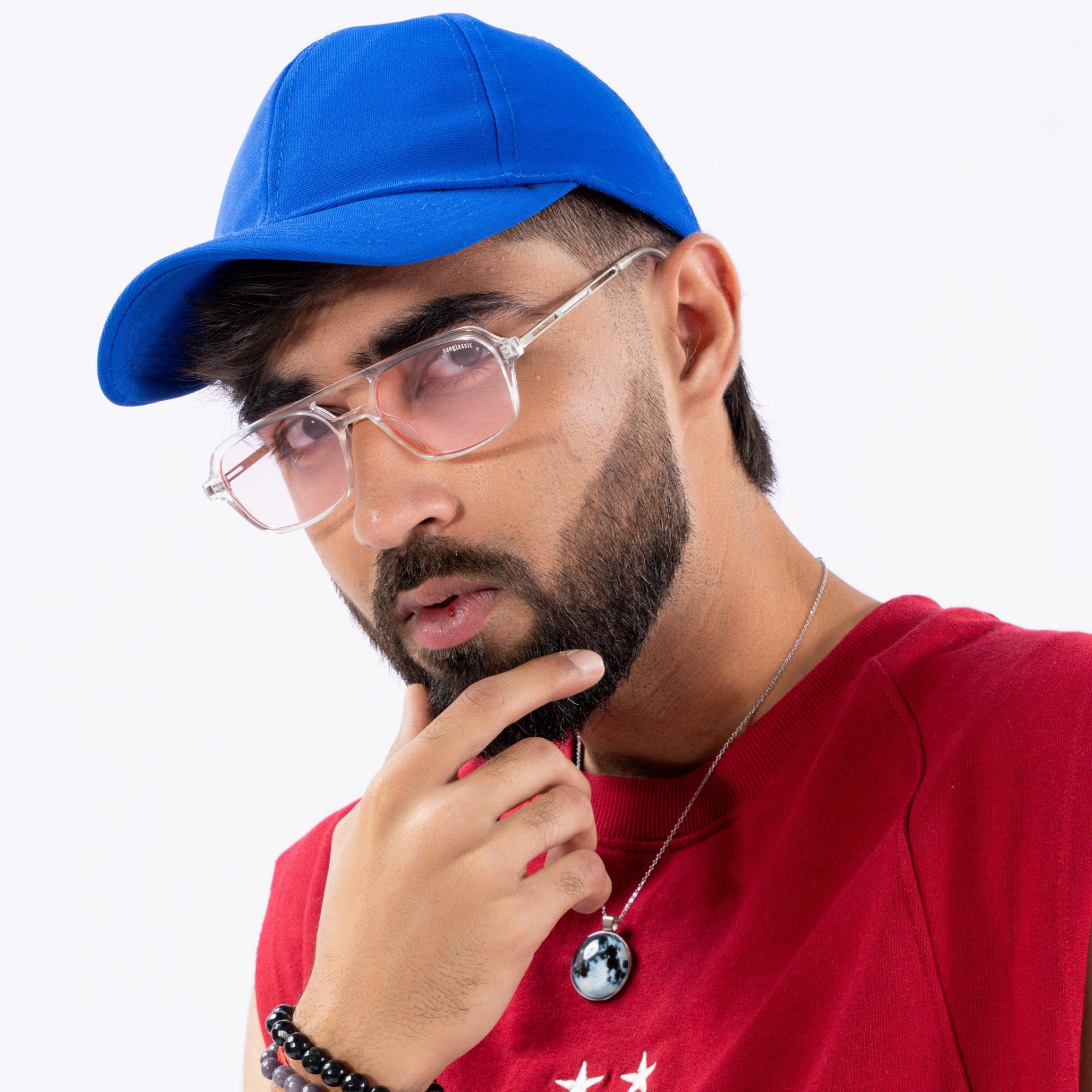 Colton Clear Pink Rectangle Sunglasses – Sunglassic.com. Male model wearing transparent-frame pink-tinted sunglasses, styled with a royal blue cap and red tee, posing thoughtfully against a white background.