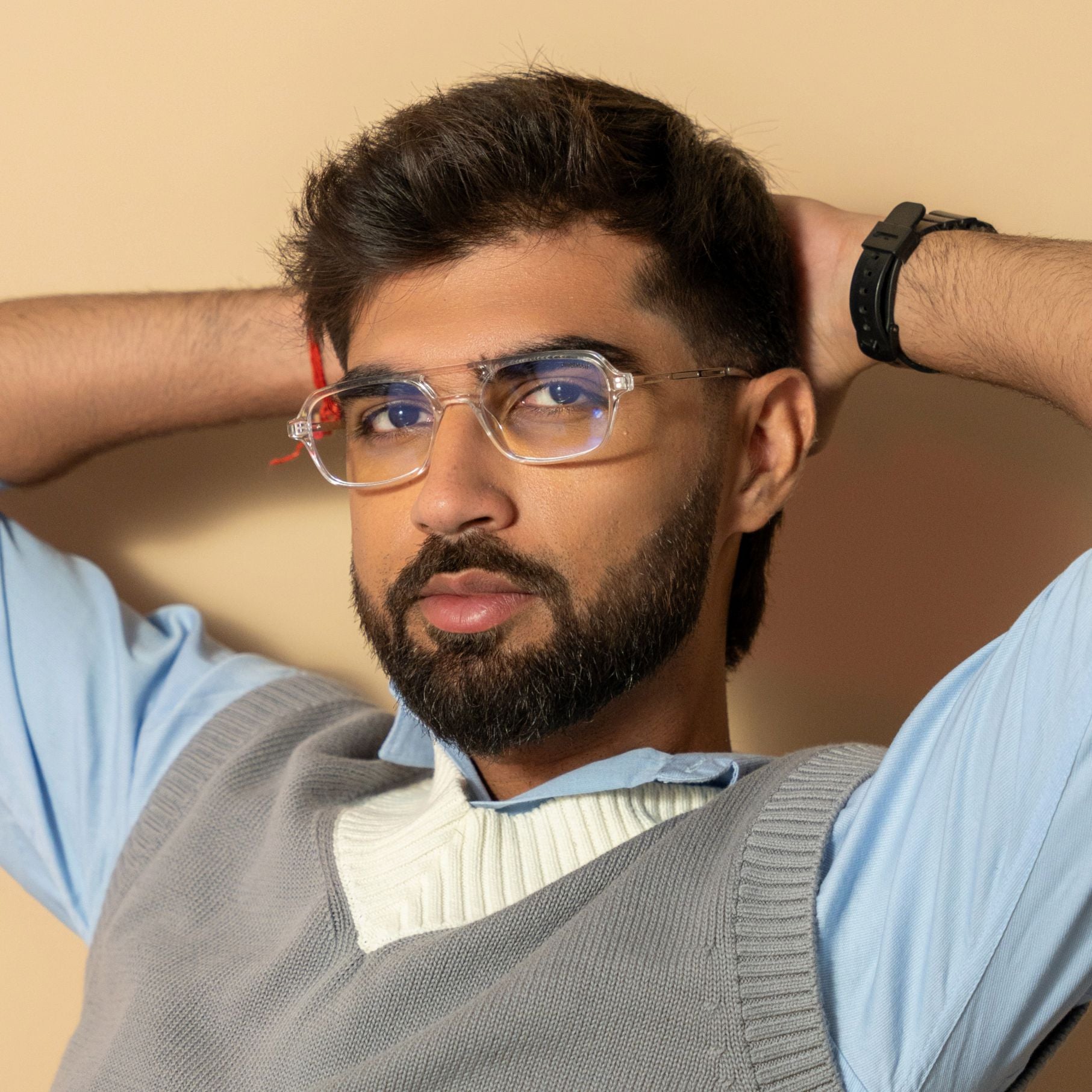 Colton Silver Clear Rectangle Sunglasses – Sunglassic.com. Male model in transparent-frame rectangle sunglasses with silver detailing and clear lenses, styled in a blue shirt with a grey sweater vest, leaning back against a beige background.