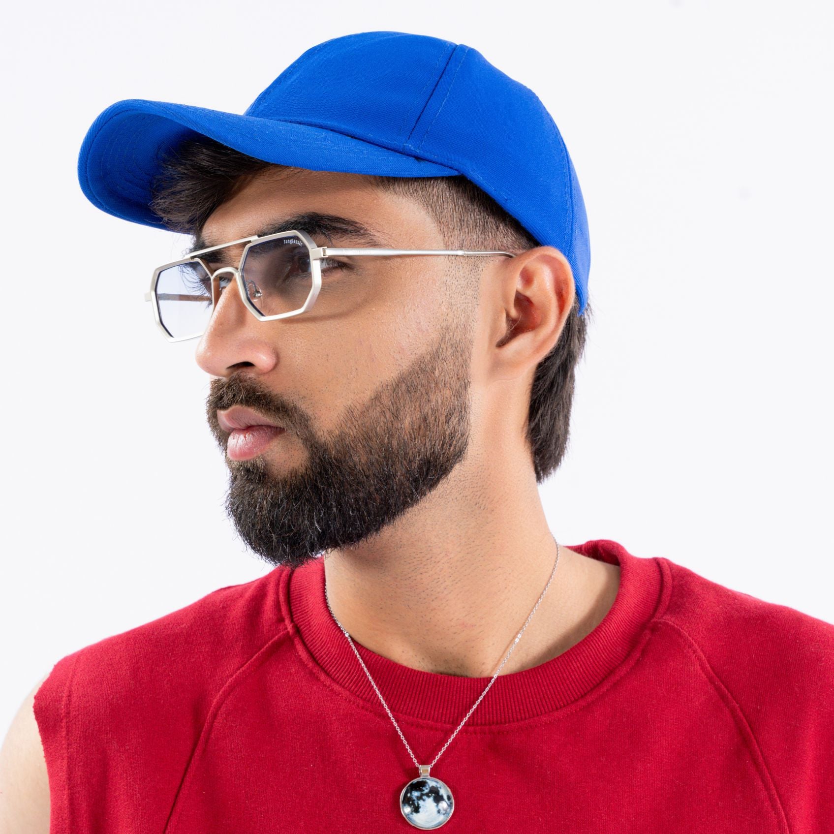 Commando V1 Rectangle Silver Clear Sunglasses – Sunglassic.com. Male model in metallic silver-rimmed rectangle sunglasses with clear lenses, paired with a blue cap and red sleeveless sweatshirt, looking sideways against a plain white background.