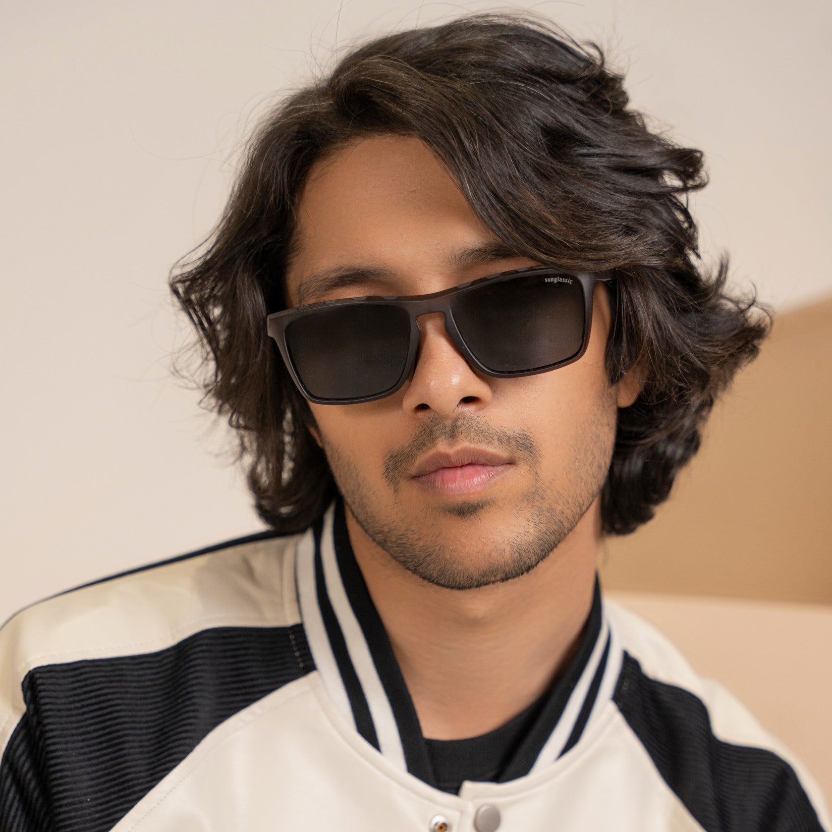 Hawk Grey Black Polarized Rectangle TR90 Sunglasses – Sunglassic.com. Male model wearing sleek polarized rectangle sunglasses with black lenses and grey TR90 frames, styled in a sporty varsity jacket, posing confidently against a neutral background.