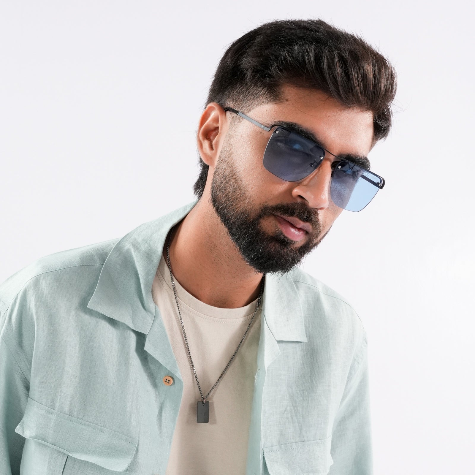 Heldish Black Blue Square Sunglasses – Sunglassic.com. Male model wearing gradient blue-tinted square sunglasses with black rims, styled in a pastel green shirt and beige tee, posing against a white background.