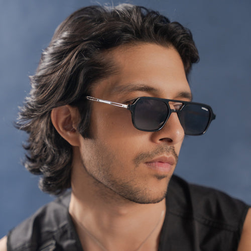 Colton Full Black Rectangle Sunglasses – Sunglassic.com. Male model wearing sleek full black rectangle sunglasses with dark lenses, styled in a sleeveless black outfit against a textured blue backdrop